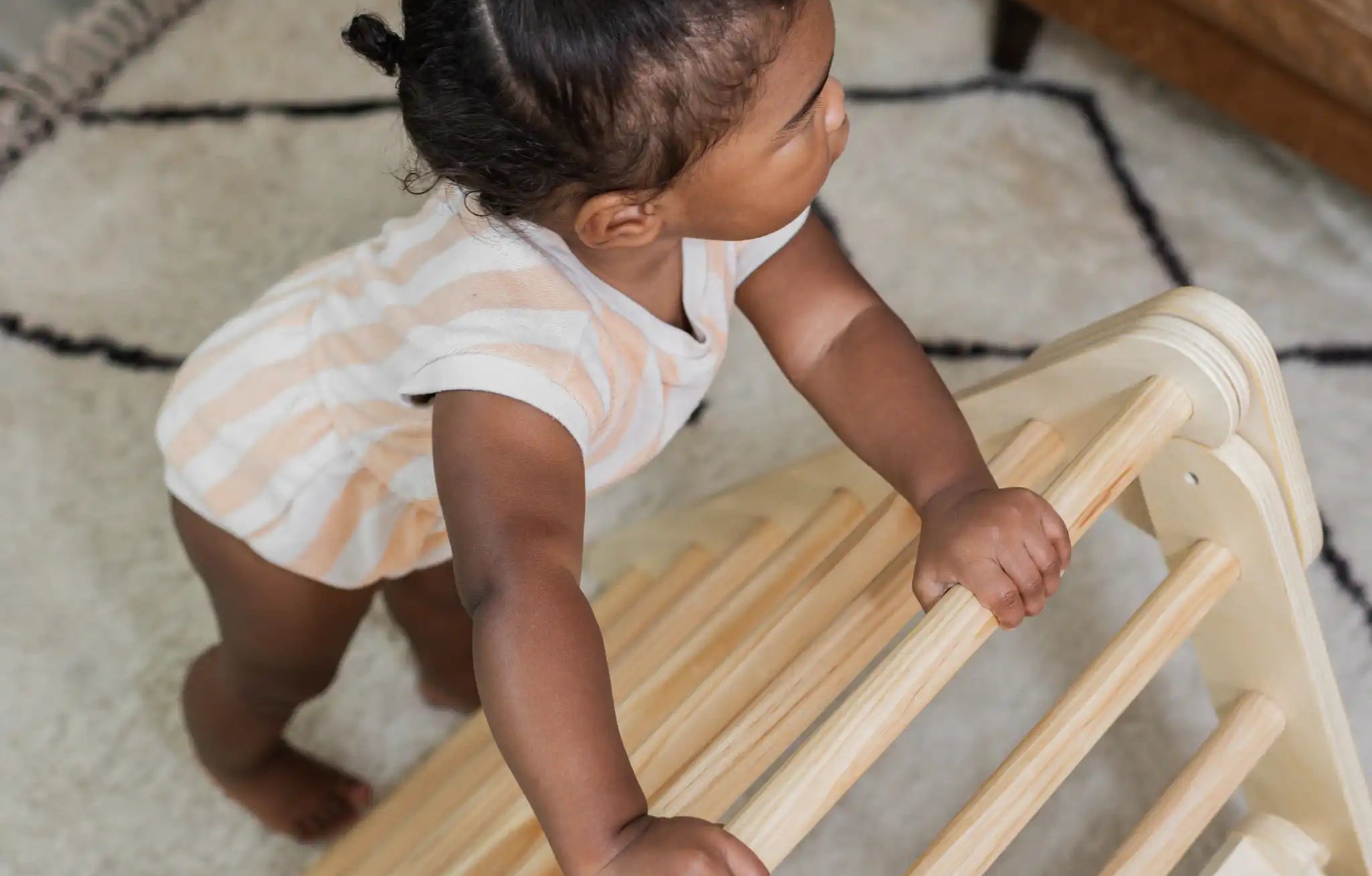 A baby pulls up to stand on the rungs of a wooden Poppyseed Play climbing triangle at home.