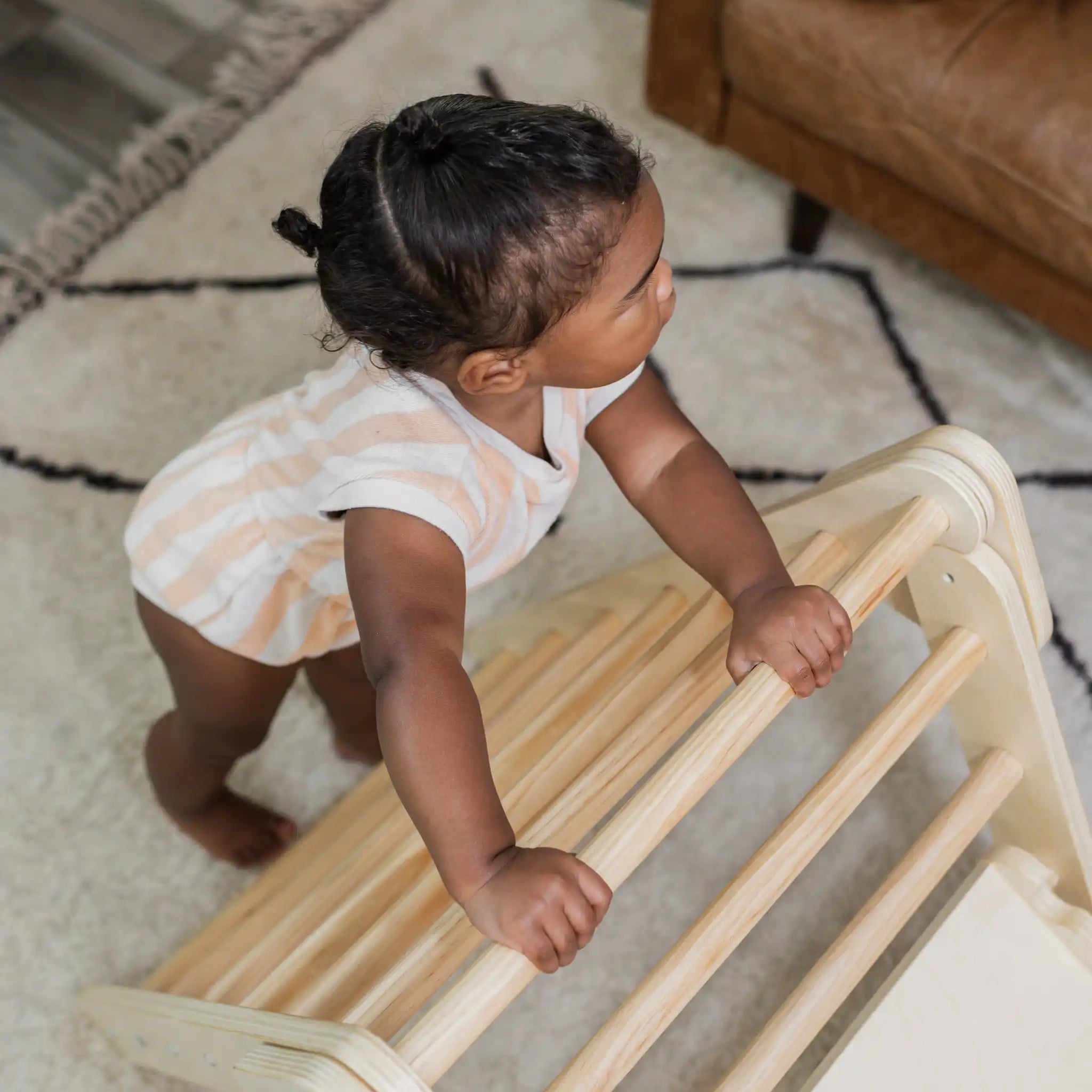A baby pulls up to stand on the rungs of a wooden Poppyseed Play climbing triangle at home.