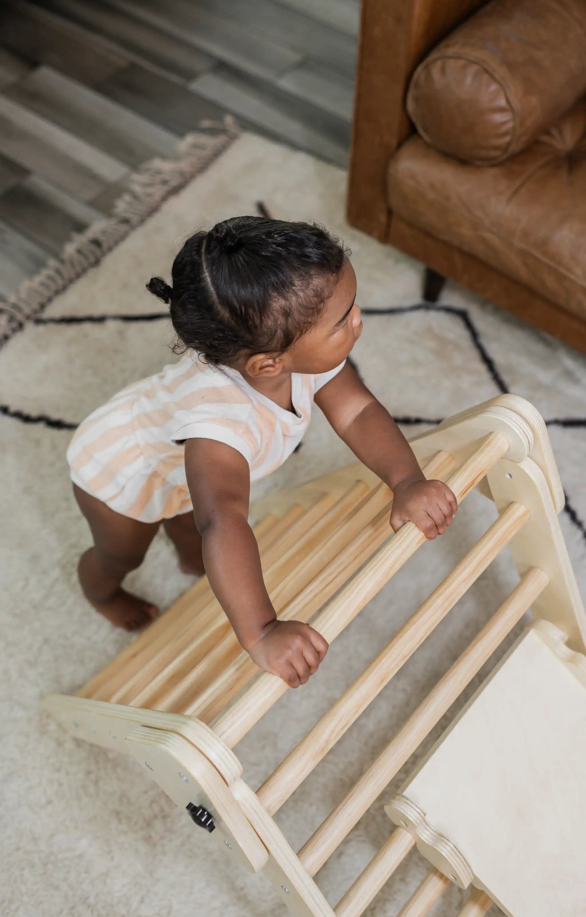 A baby pulls up to stand on the rungs of a wooden Poppyseed Play climbing triangle at home.