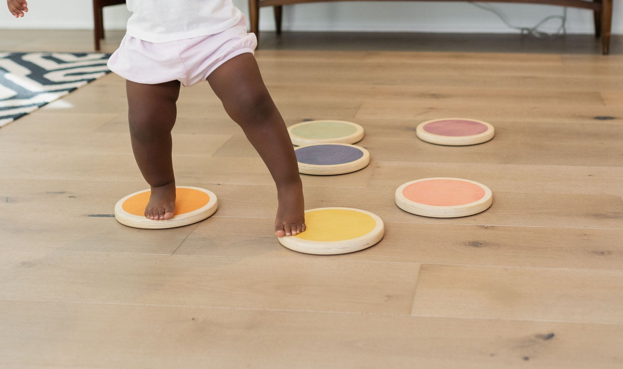 A toddler steps mid-stride between colorful wooden Poppyseed Play stepping stones across a sunlit hardwood living room.