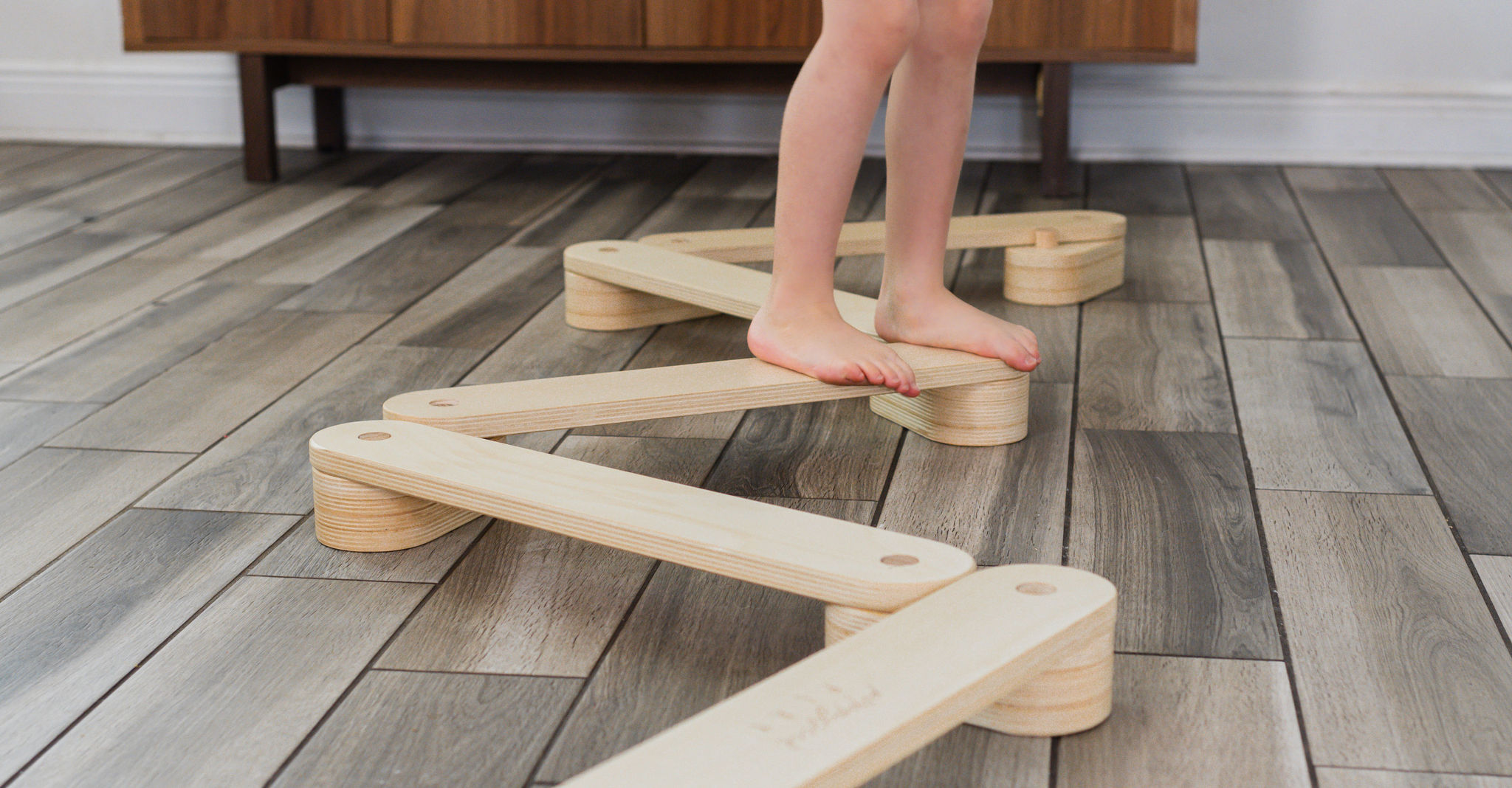 Young child walking across a wooden balance beam arranged in a zigzag pattern on the floor.