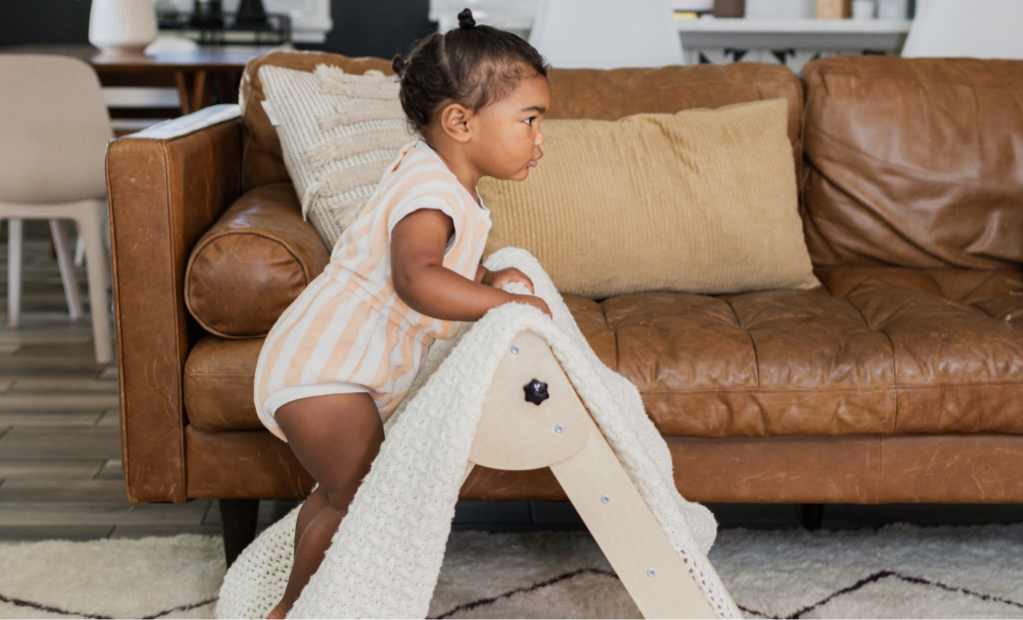 Baby in a romper climbing a Montessori Climber covered with a white knit blanket in a modern living room.