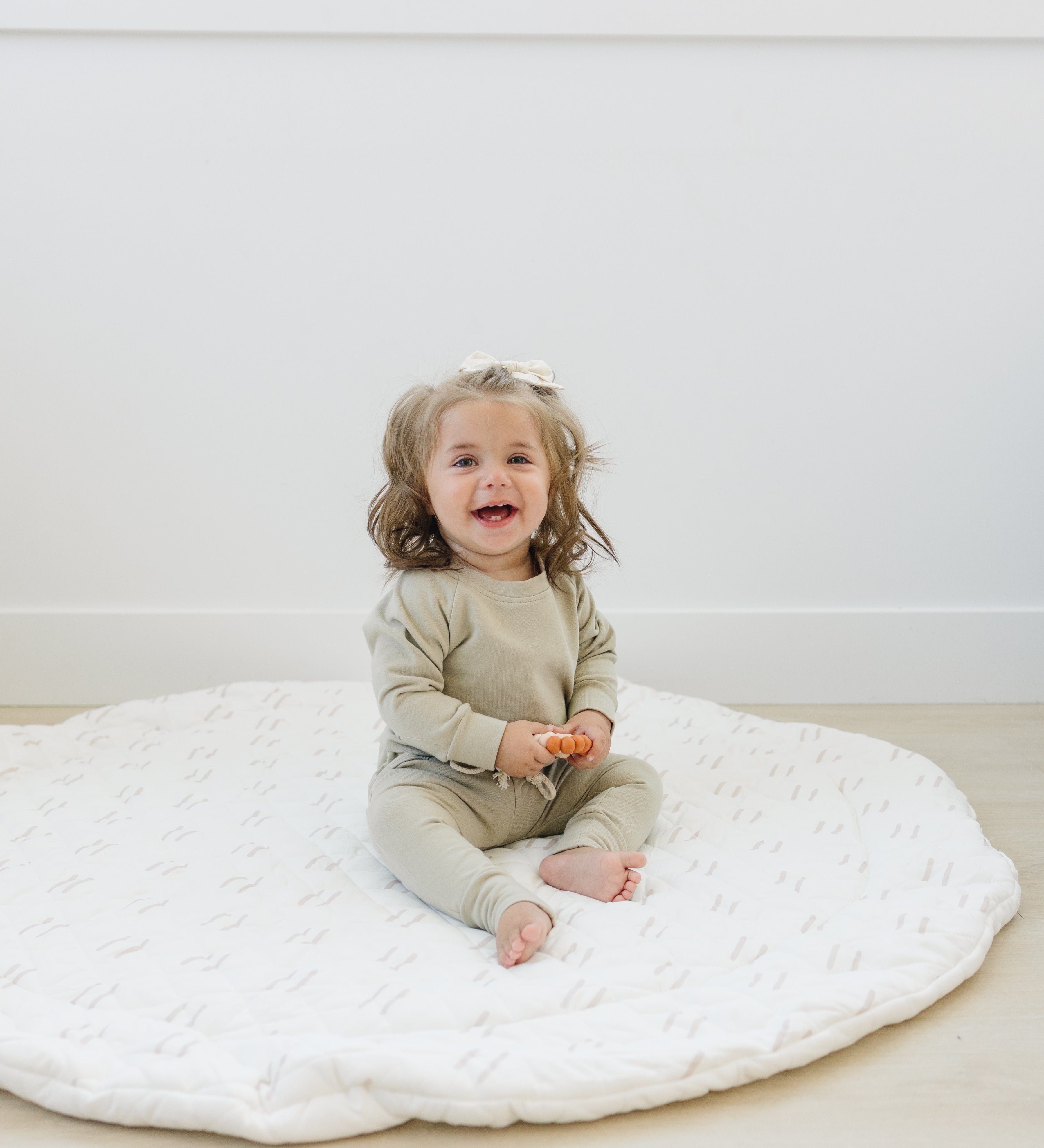 Baby sitting on a padded baby play mat, holding a toy in a bright white room.