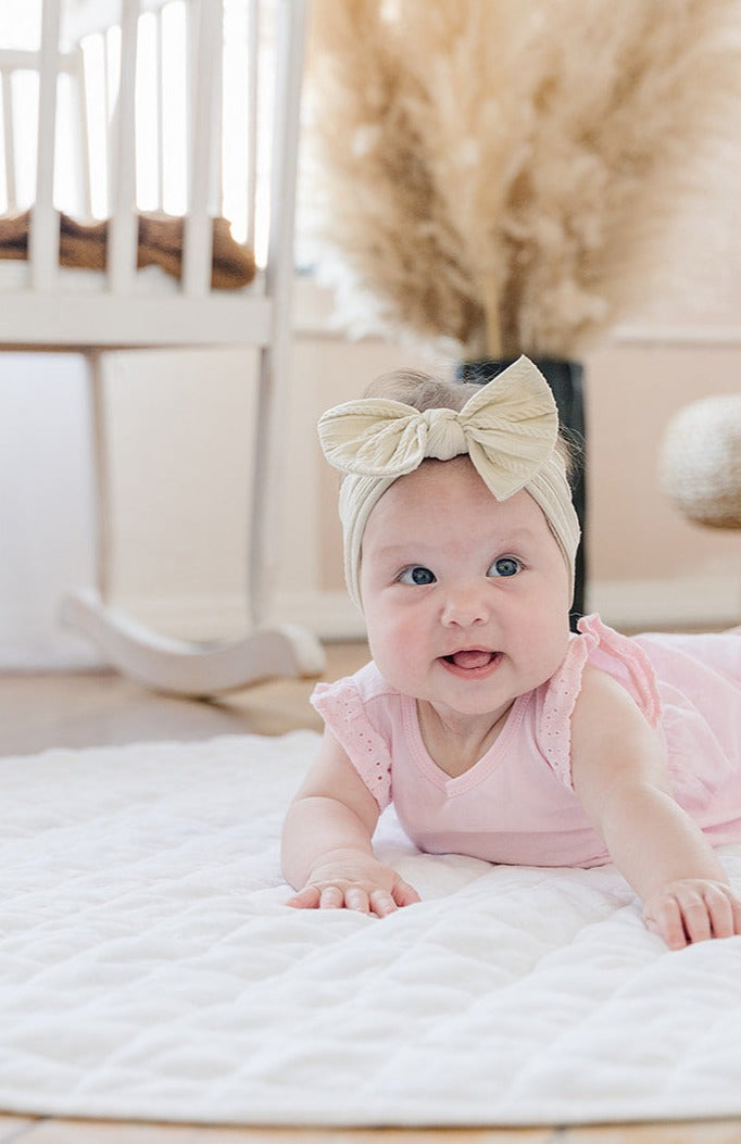 Baby in a pink outfit lying on a white linen infant play mat in a nursery.