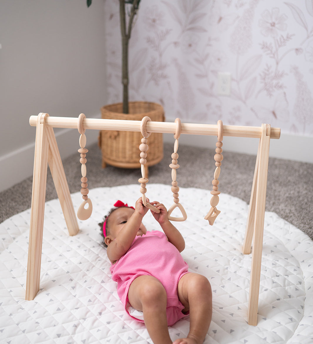Baby in pink outfit playing with a wooden play gym on a white mat.