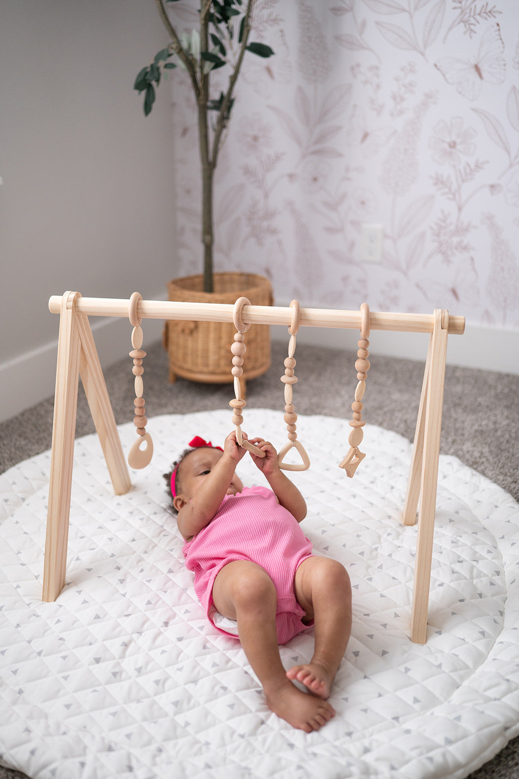 Baby in pink outfit playing with a wooden play gym on a white mat.
