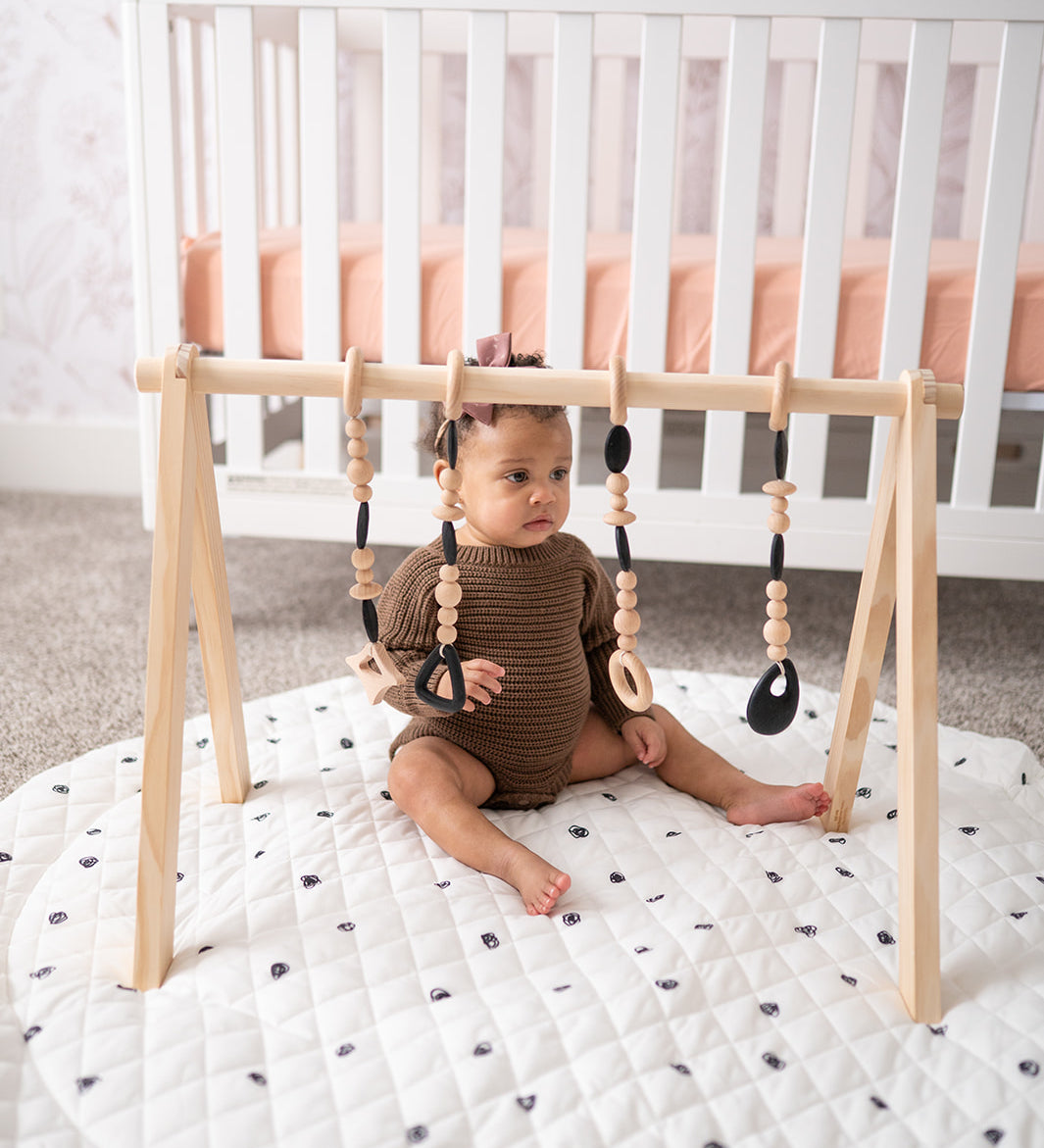 Baby sitting on a white mat with a wooden baby gym in a nursery.