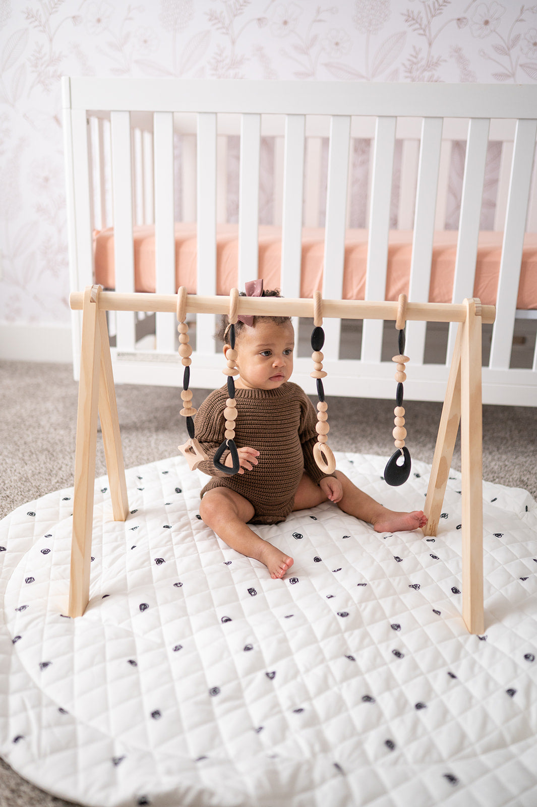 Baby sitting on a white mat with a wooden baby gym in a nursery.