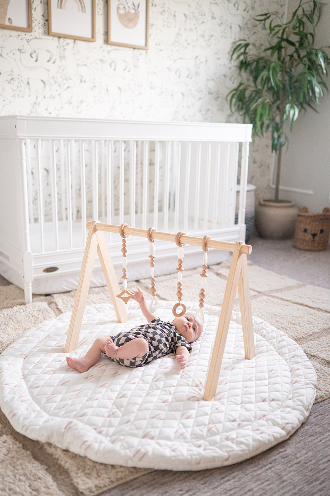 Baby playing on a white mat with a wooden play gym in a nursery.