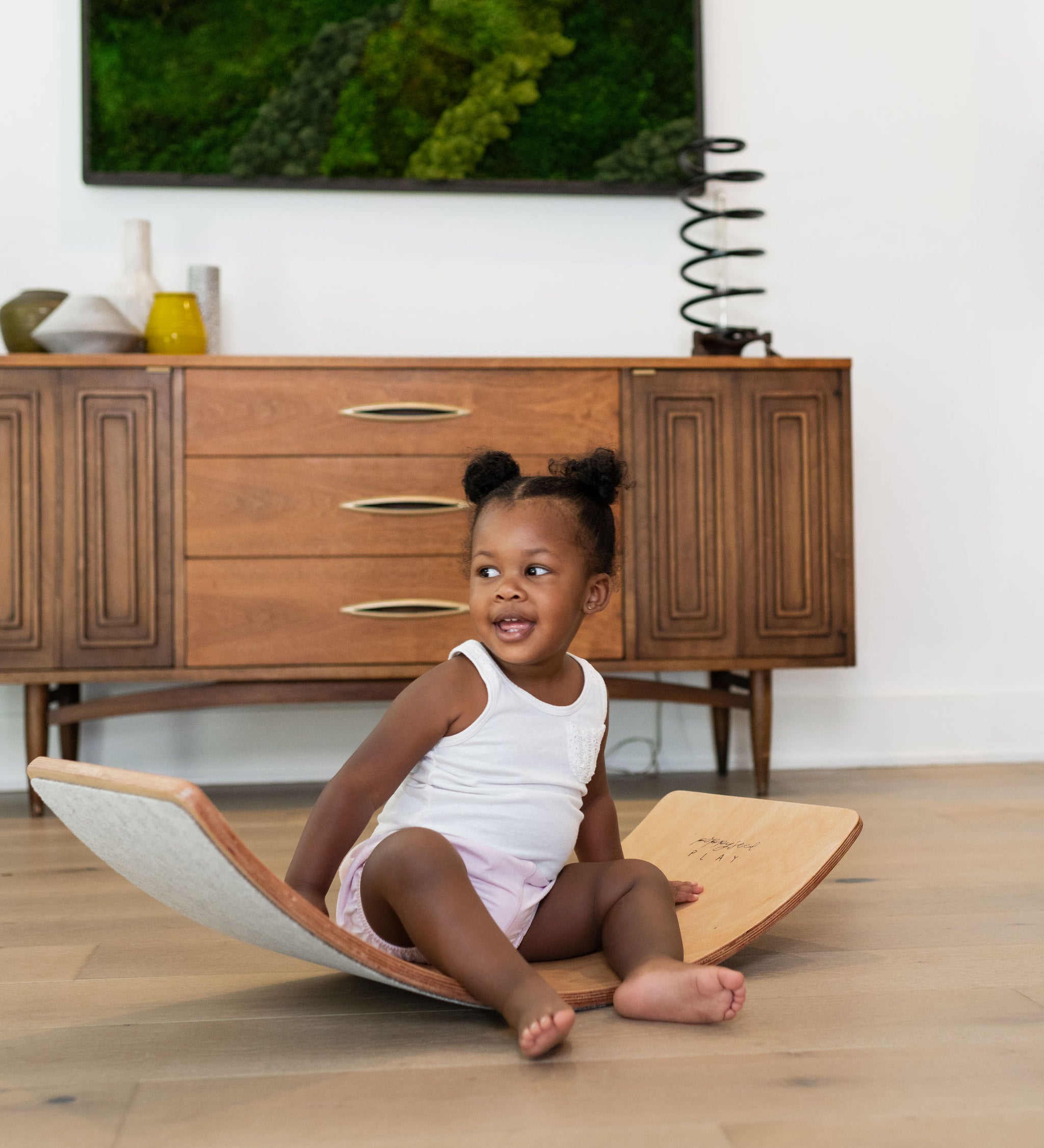 Toddler sitting and playing on a wooden balance board in a modern living room.