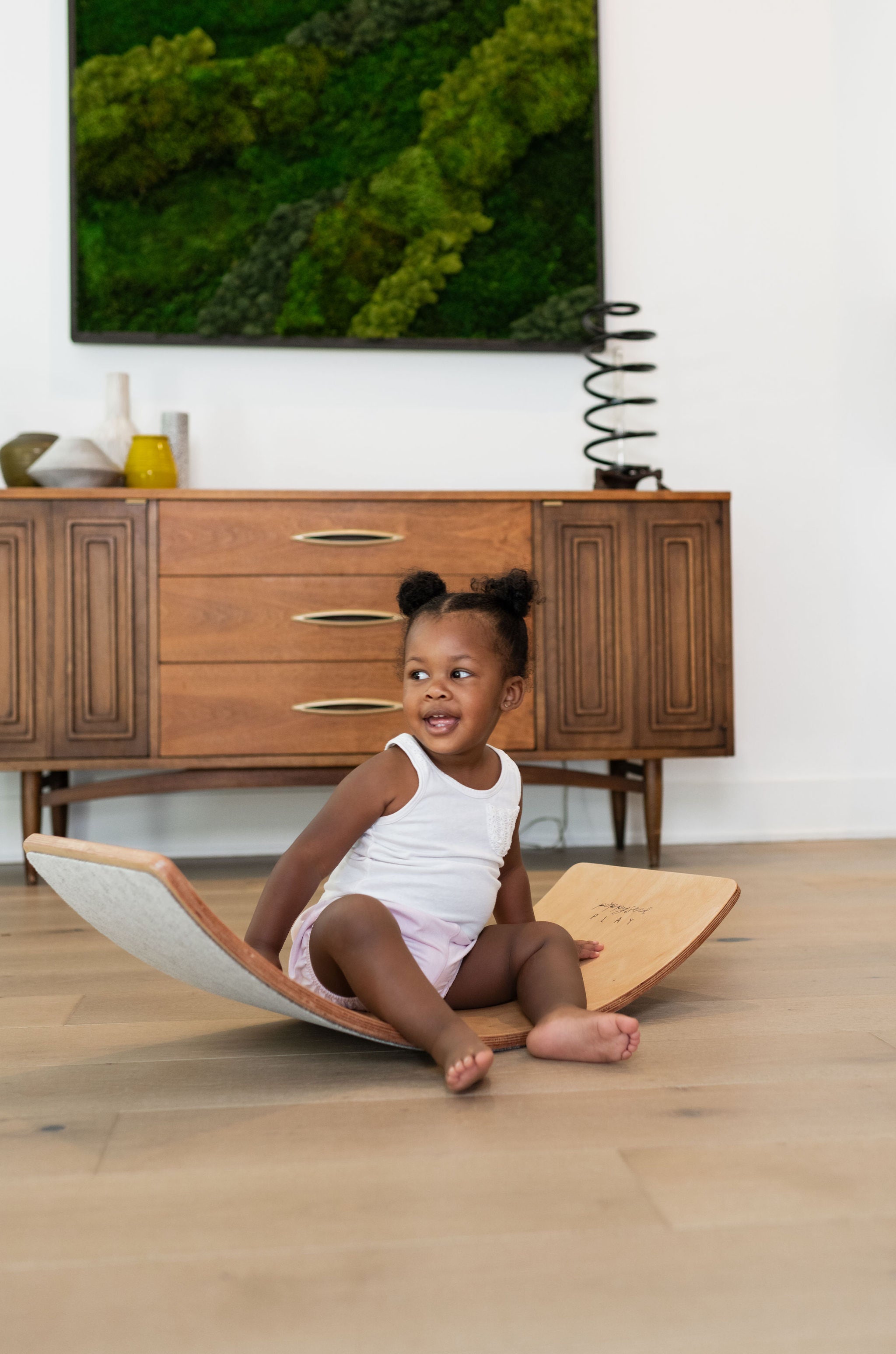 Toddler sitting and playing on a wooden balance board in a modern living room.