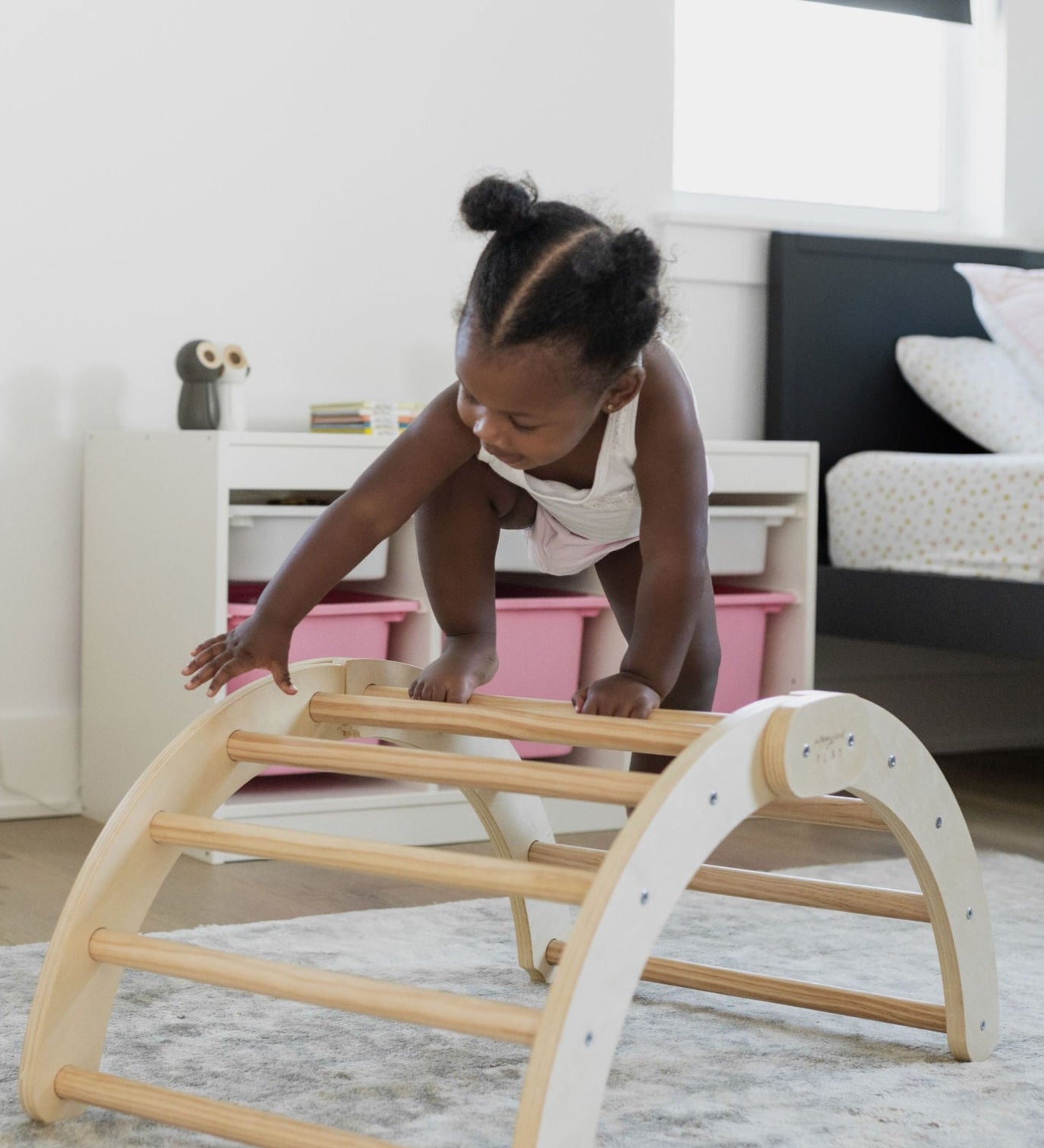 Toddler climbing over a Poppyseed Play wooden foldable Climbing Arch & Rocker on a soft rug.