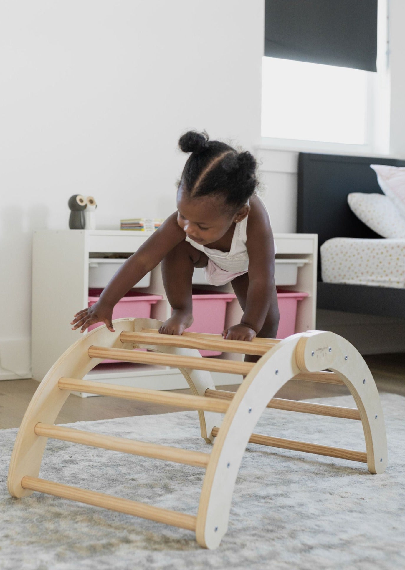 Toddler climbing over a Poppyseed Play wooden foldable Climbing Arch & Rocker on a soft rug.