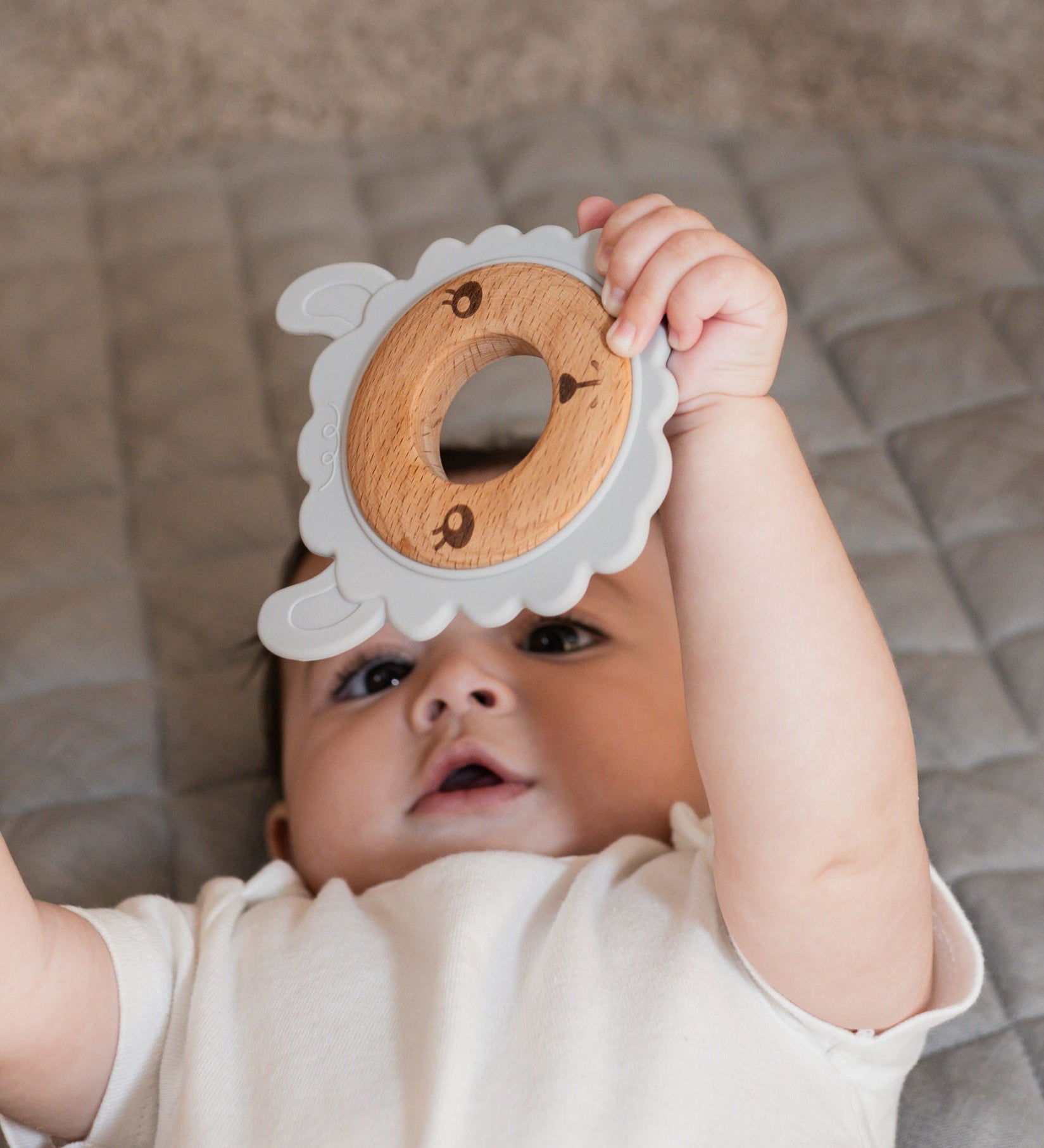 Baby lying on a play mat holding a silicone & wood teether while looking upward.