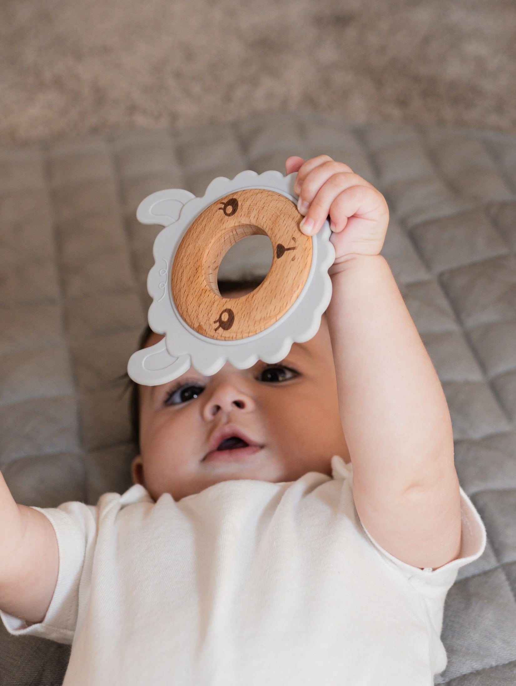 Baby lying on a play mat holding a silicone & wood teether while looking upward.