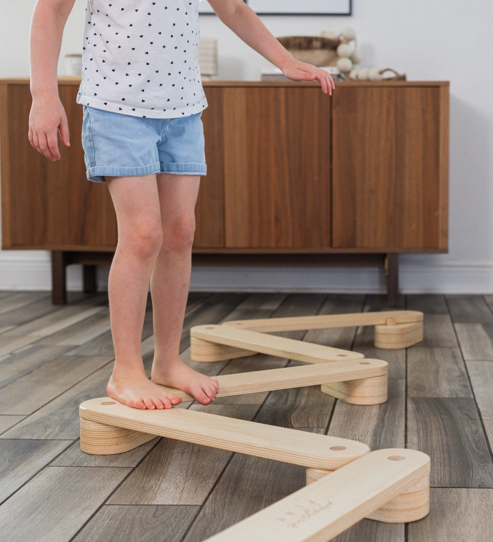 Young child balancing and walking along a wooden balance beam over a wood floor.