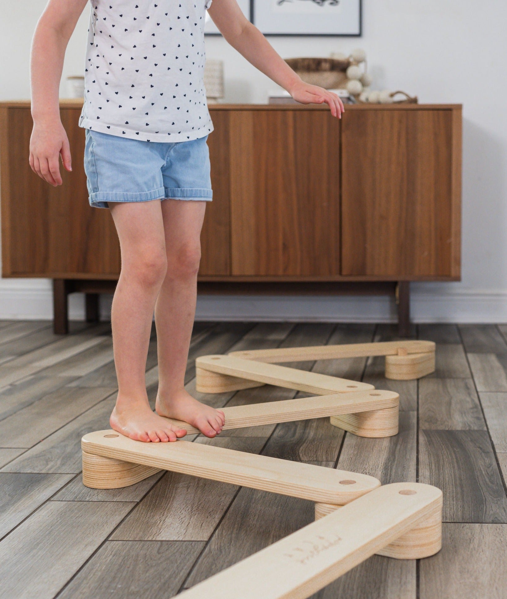 Young child balancing and walking along a wooden balance beam over a wood floor.