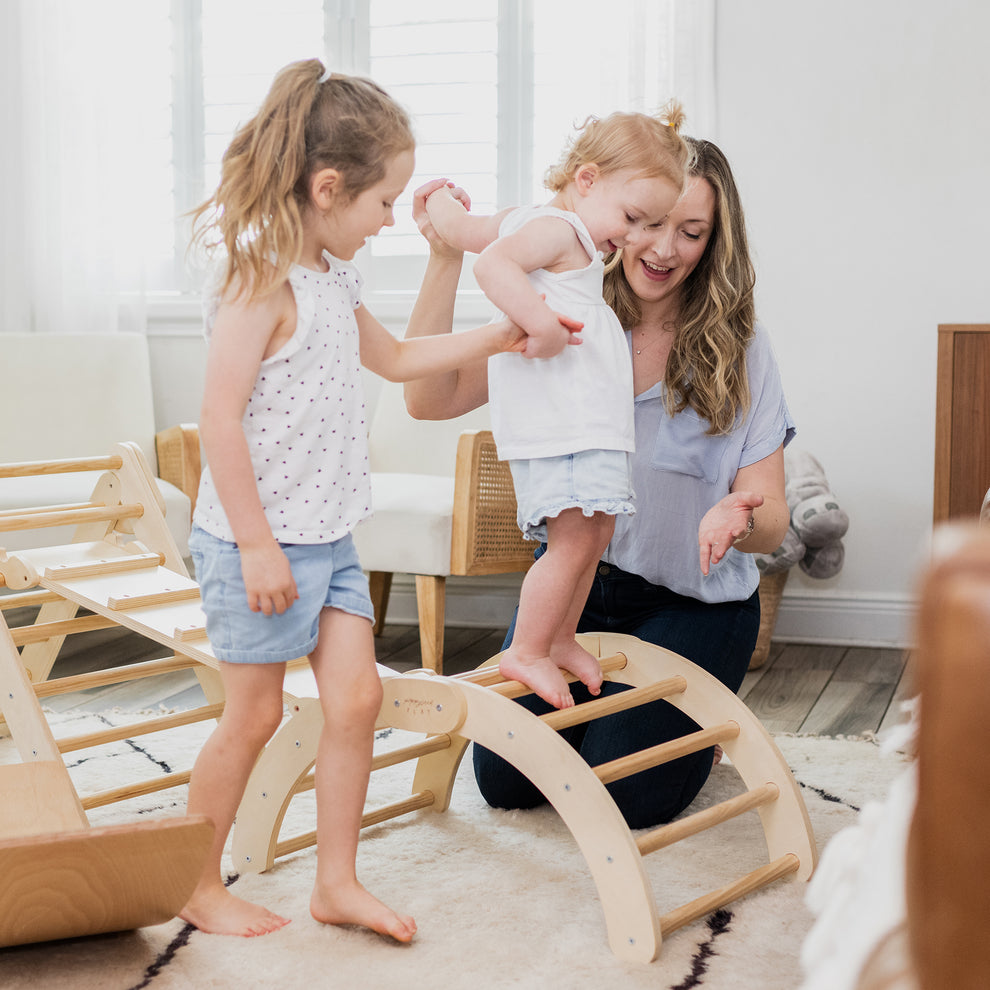 Woman helping a toddler walk across a climbing arch while another young child stands nearby in a living room.