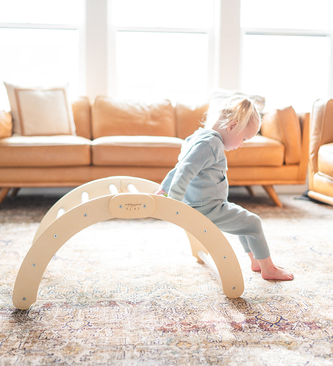 A child playing on a beige wooden climbing arch indoors, with furniture in the background.