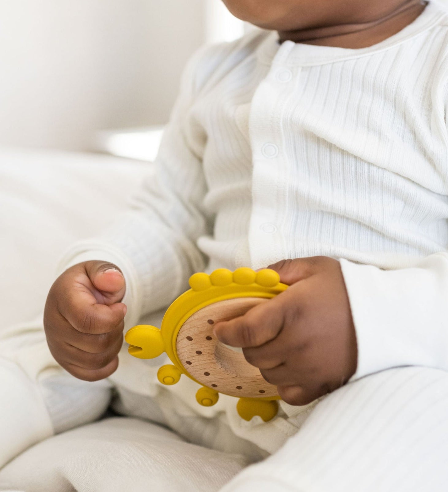 Baby in a white ribbed sleeper holding a yellow silicone & wood teether.
