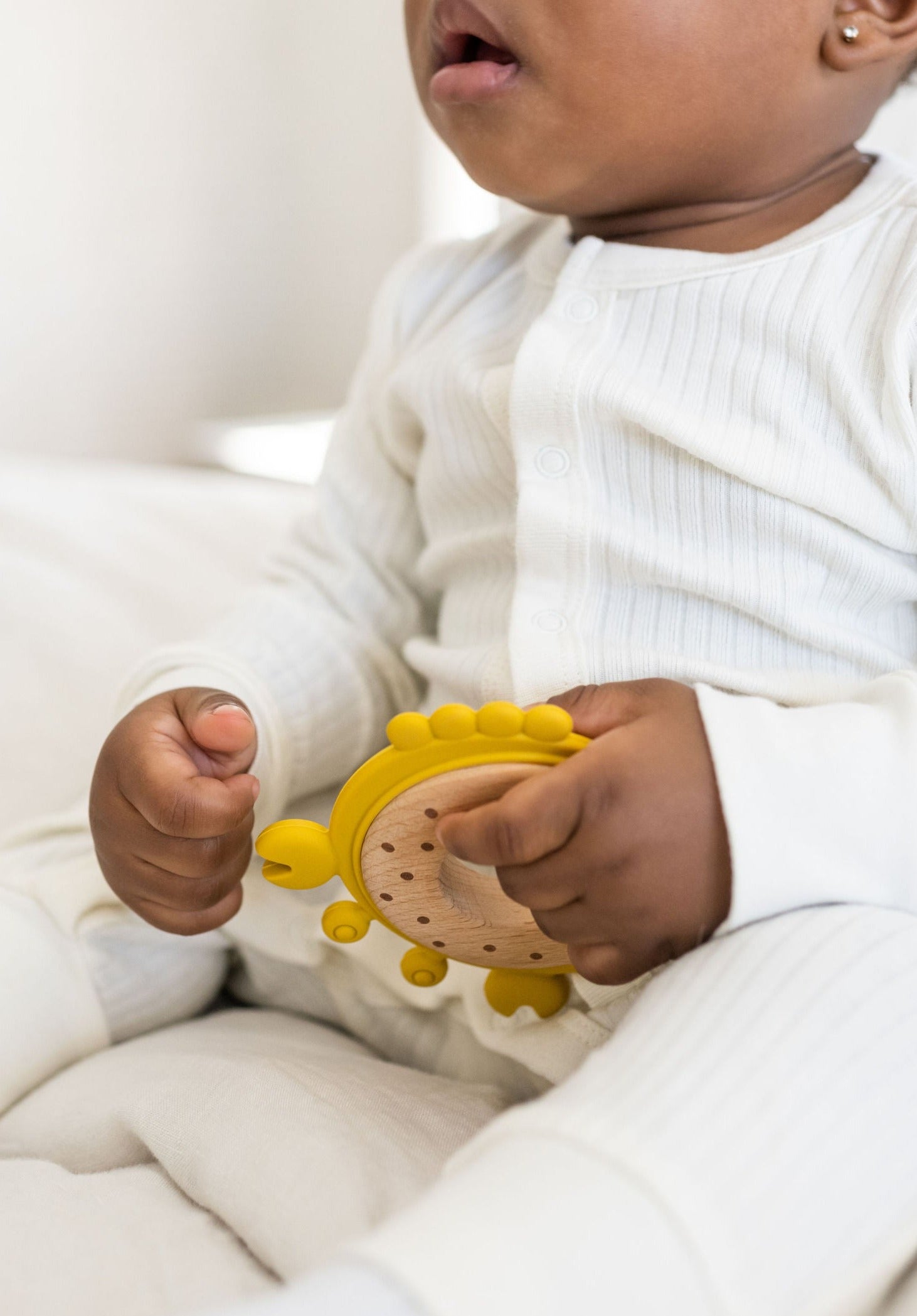 Baby in a white ribbed sleeper holding a yellow silicone & wood teether.