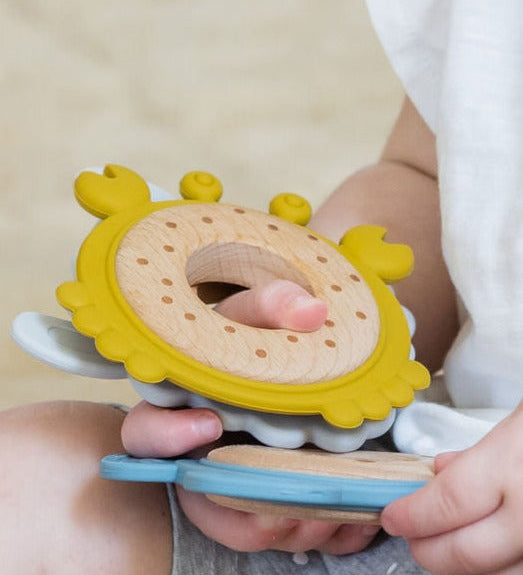 Baby holding a Teether Trio Bundle featuring yellow, blue, and grey silicone & wood teethers.