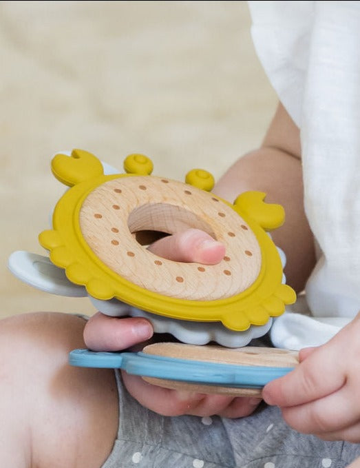 Baby holding a Teether Trio Bundle featuring yellow, blue, and grey silicone & wood teethers.