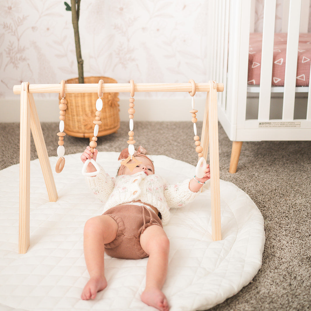Baby lying on a padded baby play mat reaching for wooden toys on a wooden baby gym in a bright nursery.