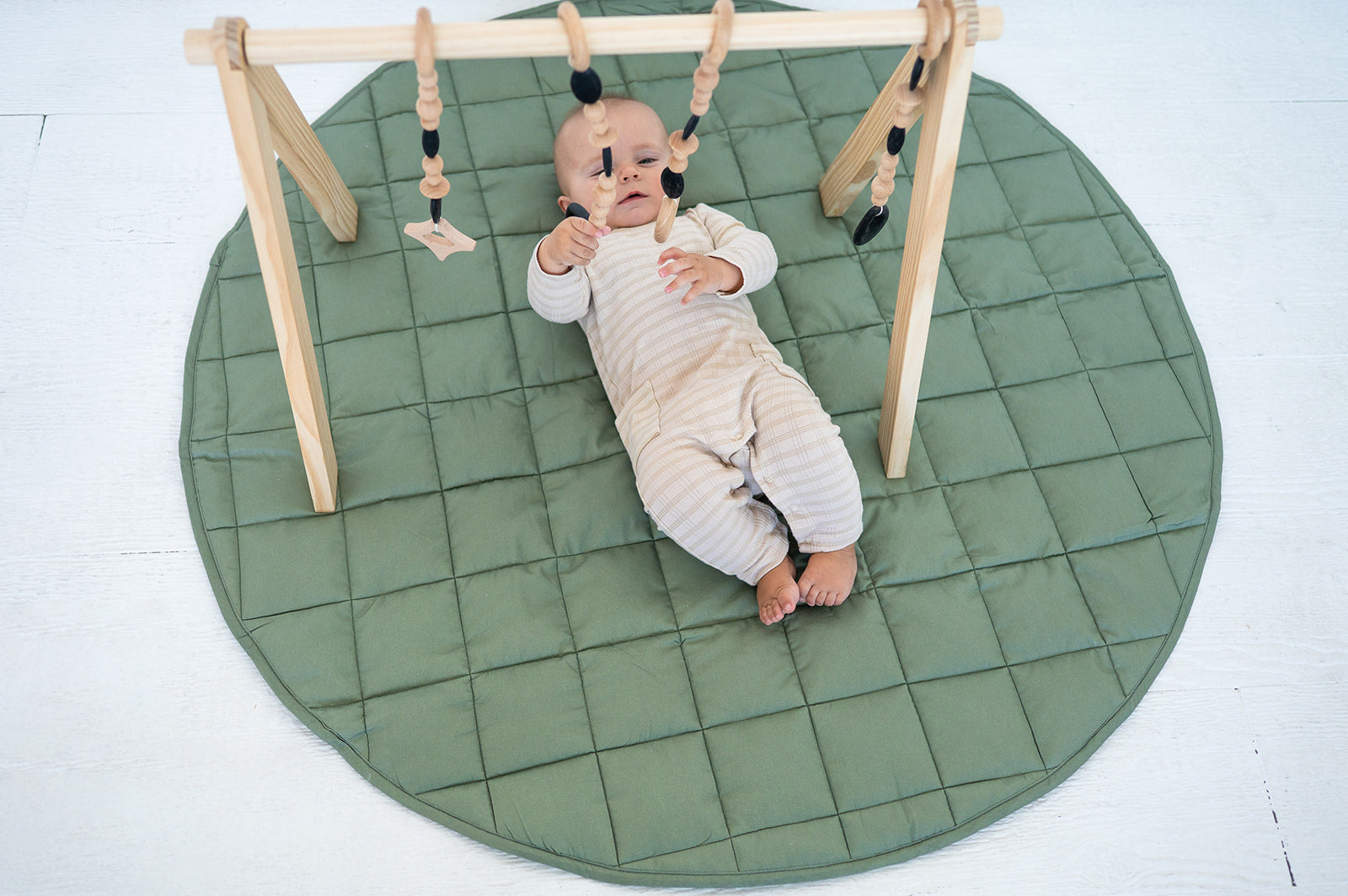 Baby under a wooden baby gym on the olive Neutral Round Baby Play Mat, seen from above