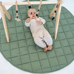Baby under a wooden baby gym on the olive Neutral Round Baby Play Mat, seen from above