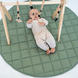 Baby under a wooden baby gym on the olive Neutral Round Baby Play Mat, seen from above
